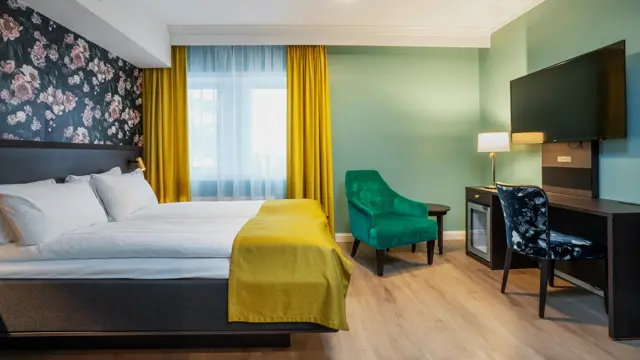 Turkise vegger, mørkt skrivebord med veggehengt TV, og mønstrete stol. Grønn lenestol. Vindu med gul gardin. Dobbeltseng med gult sengeteppe. I dobbeltrom på Thon Hotel Høyers i Skien.