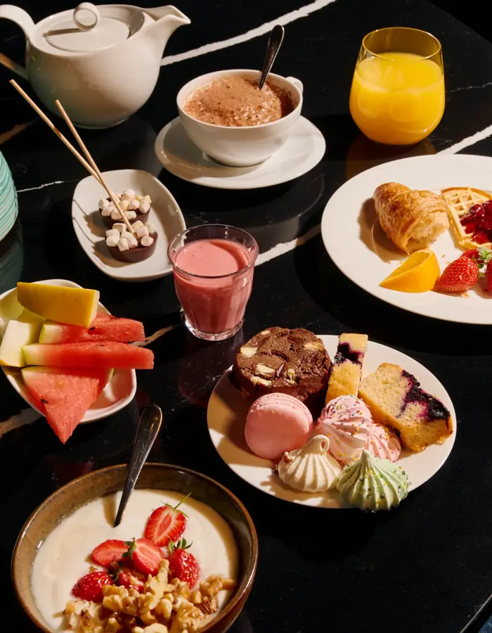Et frokostbord med yoghurt, en tallerken med småkaker og frukt. Det er også kakao, en tekanne, smoothie, appelsinjuice og en tallerken med croissant, vaffel og syltetøy