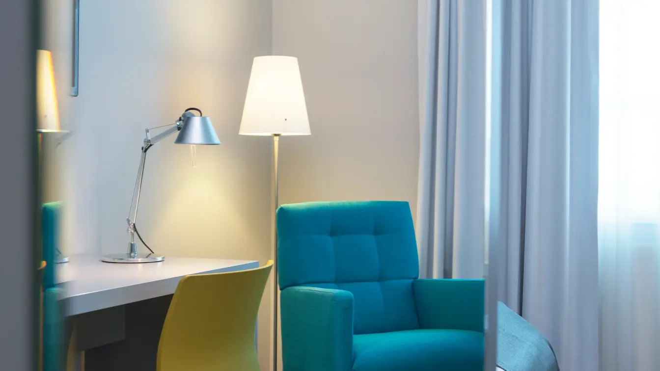 Nærbilde av Standard Single Room på Thon Hotel Trondheim. Turkis lenestol, hvitt skrivebord, gul kontorstol, leselampe, stålampe og hvite gardiner.