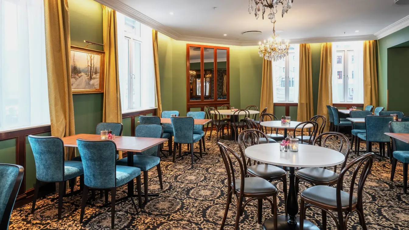 Turkise vegger, vinduer med gule gardiner fra tak til gulv. Store lysekroner. Mørke kvadratiske bord, blå stoler og mørkt mønstrete gulvteppe. I restauranten på Thon Hotel Høyers i Skien. 