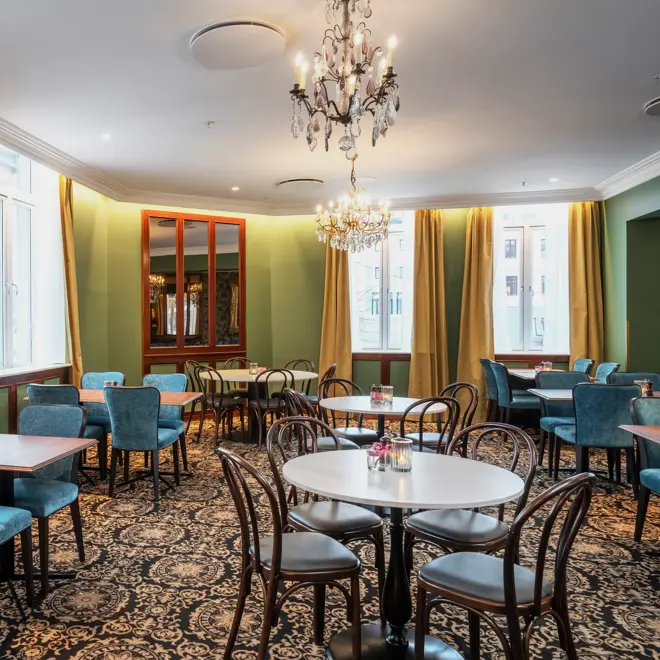 Turkise vegger, vinduer med gule gardiner fra tak til gulv. Store lysekroner. Mørke kvadratiske bord, blå stoler og mørkt mønstrete gulvteppe. I restauranten på Thon Hotel Høyers i Skien. 