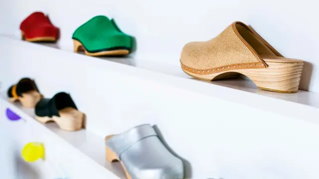 Utvalg av tresko i ulike farger på hvit hylle på hvit vegg. Illustrasjonsbilde til artikkel om vårmote for sko.
