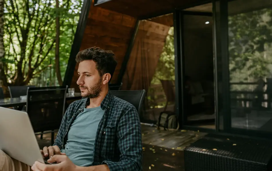 Mann med rutete skjorte sitter med laptop i fanget utenfor hytte