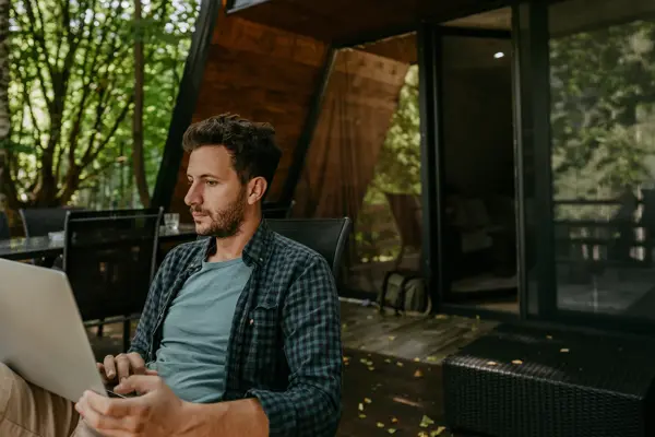 Mann med rutete skjorte sitter med laptop i fanget utenfor hytte