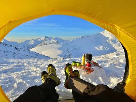 To par bein som kommer ut av en gul teltåpning i retning snødekte topper og leirplass.