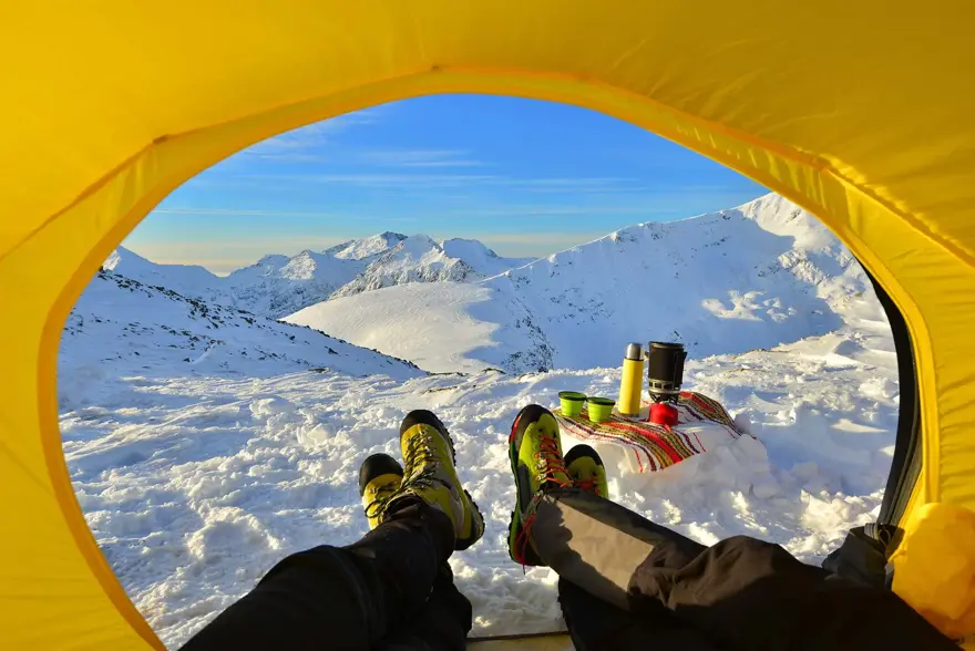 To par bein som kommer ut av en gul teltåpning i retning snødekte topper og leirplass.