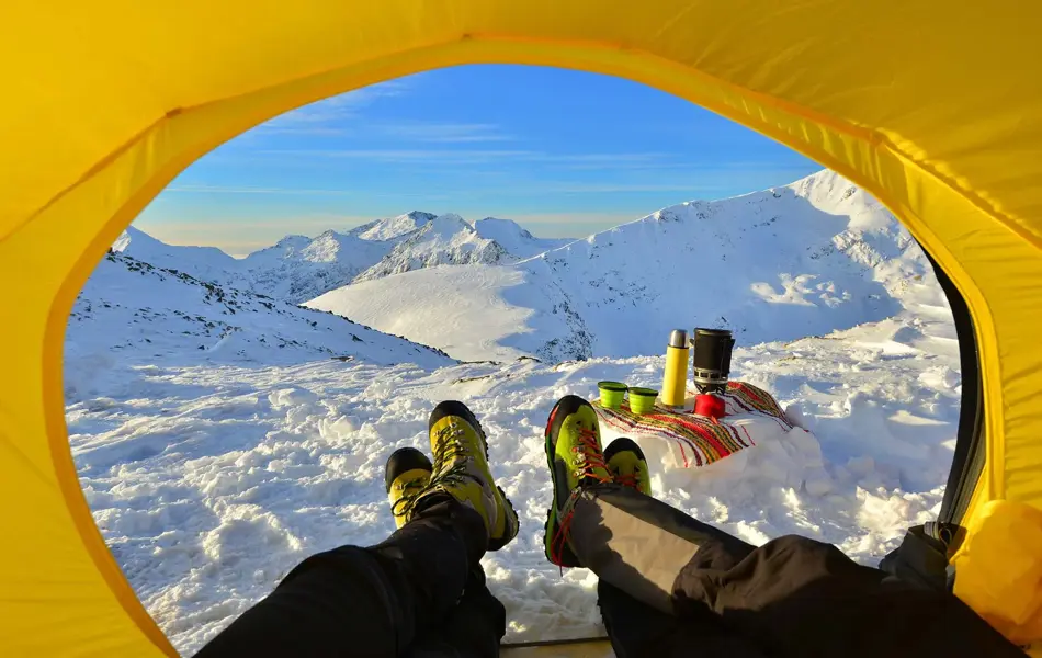 To par bein som kommer ut av en gul teltåpning i retning snødekte topper og leirplass.