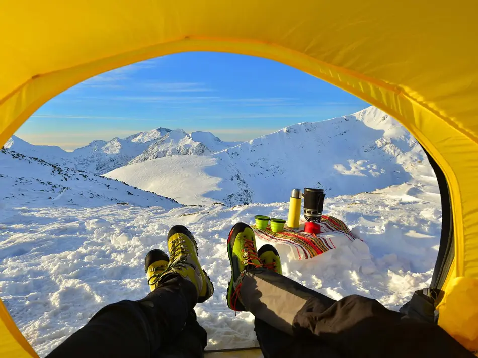 To par bein som kommer ut av en gul teltåpning i retning snødekte topper og leirplass.