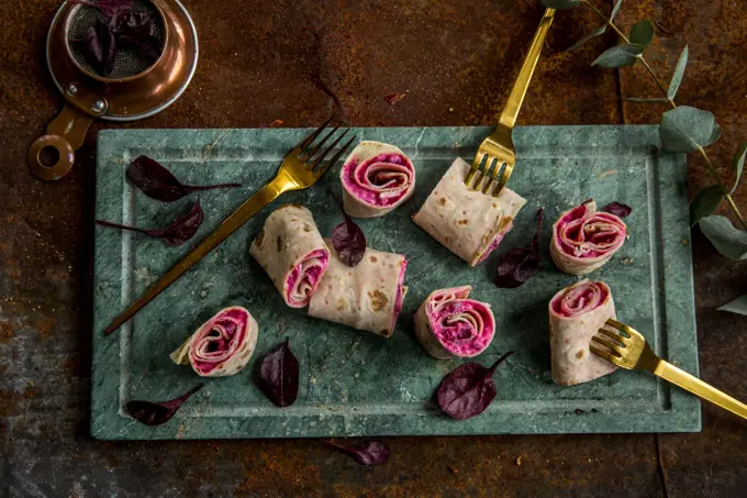 Lefseruller med rødbeter og lammerull sett ovenfra på et grønt marmorfat og gullgafler dandert rundt. 