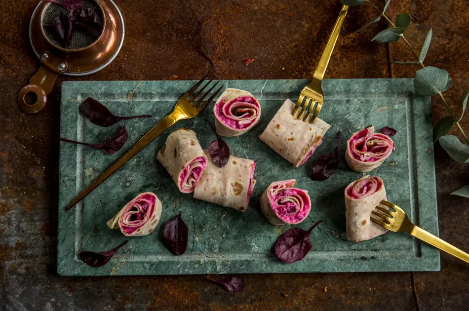 Lefseruller med rødbeter og lammerull sett ovenfra på et grønt marmorfat og gullgafler dandert rundt. 