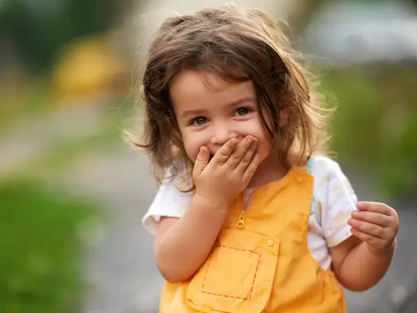 Little girl with brown hair outdoors, covering her mouth laughing