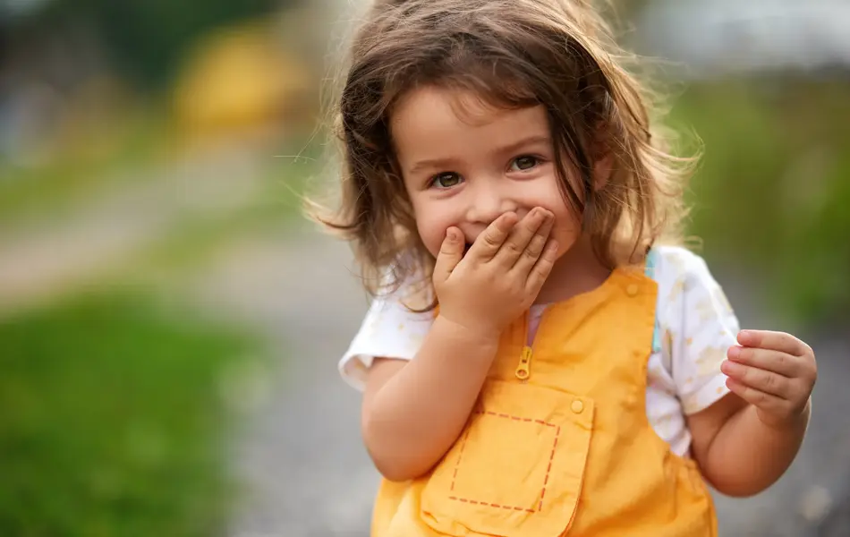 Little girl with brown hair outdoors, covering her mouth laughing