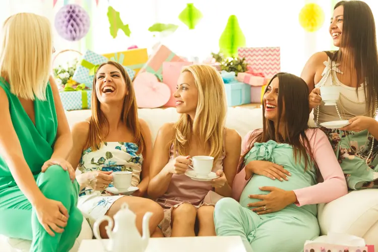 Group of female friends sitting on sofa, drinking tea on baby shower party, laughing.