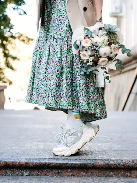 Halvportrett av dame i blomsterkjole og sneakers som holder en blomsterbukett