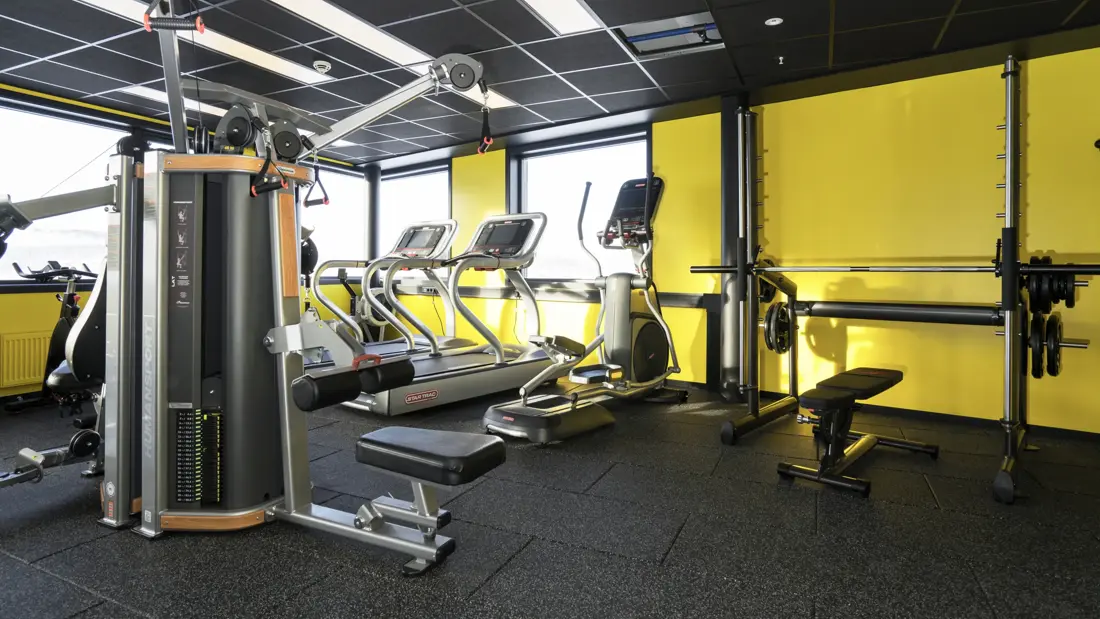 Treningsrom med forskjellige treningsapparater, gul vegg og utsikt fra vinduet på Thon Hotel Kirkenes ved kaia i Kirkenes sentrum
