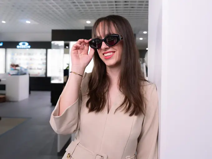 Jente med brunt halvlangt hår som står og lener seg inntil en vegg med beige genser og sorte solbriller fra Botega Veneta