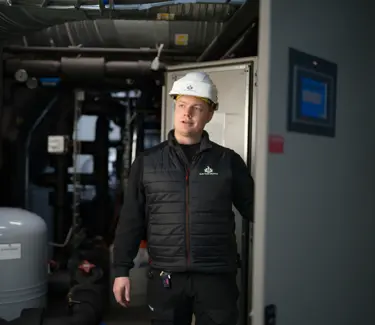 En mann står i et maskinrom. Han  har en hvit hjelm på hodet. 