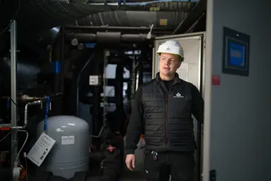 En mann står i et maskinrom. Han  har en hvit hjelm på hodet. 