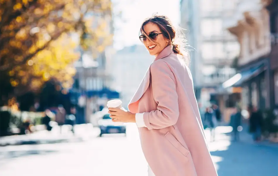 Dame smiler iført lys rosa kåpe og solbriller på gata med kaffe i hånda.