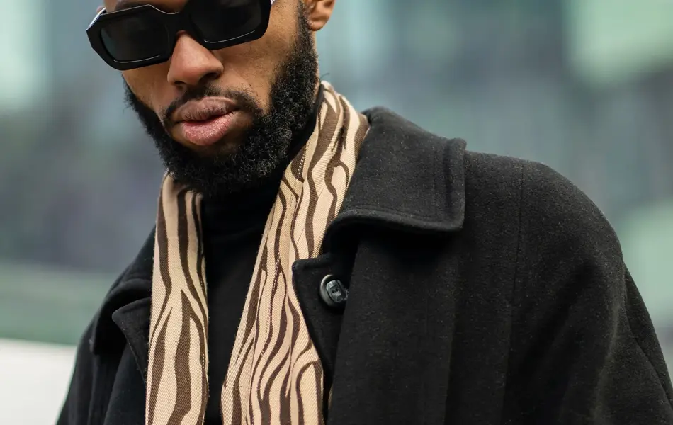 Nærbilde av en mann med oransje lue og solbriller utendørs. Mannen har svart jakke og et skjerf i halsen.