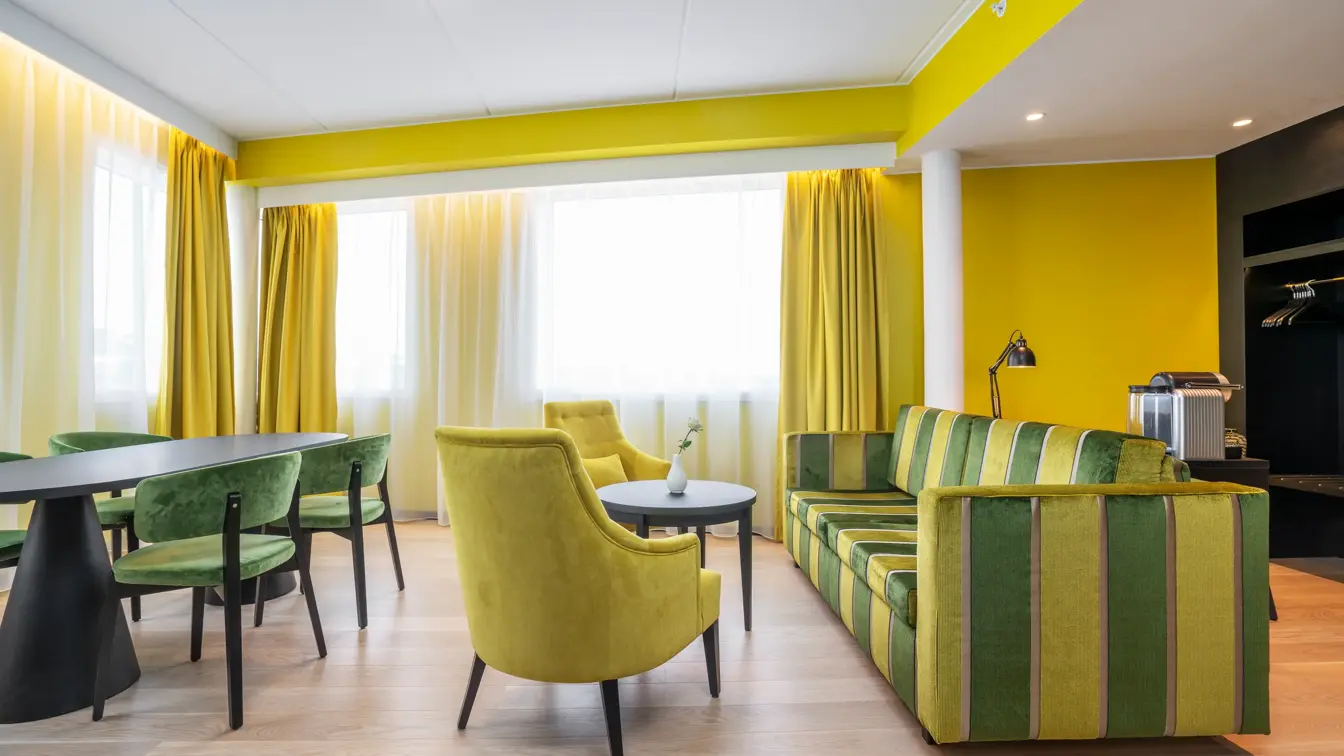 Sittegruppe med gul og grønn stripete sofa, rundt bord, gule stoler, spisegruppe med mørkt rundt bord og grønne stoler, gule vegger og gule gardiner i suite på Thon Hotel Kirkenes