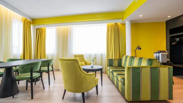 Sittegruppe med gul og grønn stripete sofa, rundt bord, gule stoler, spisegruppe med mørkt rundt bord og grønne stoler, gule vegger og gule gardiner i suite på Thon Hotel Kirkenes