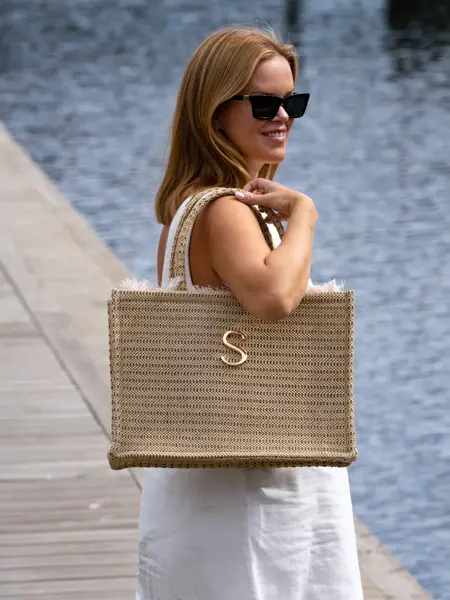 Dame står på brygge med hvit kjole, sorte solbriller og en vevd veske over skulderen.