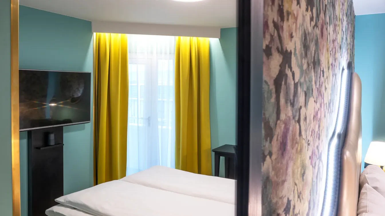 Blomstret tapet og dobbeltseng i dobbeltrom med speilbilde av resten av rommet, som har gule gardiner og tv på Thon Hotel Cecil i Oslo sentrum