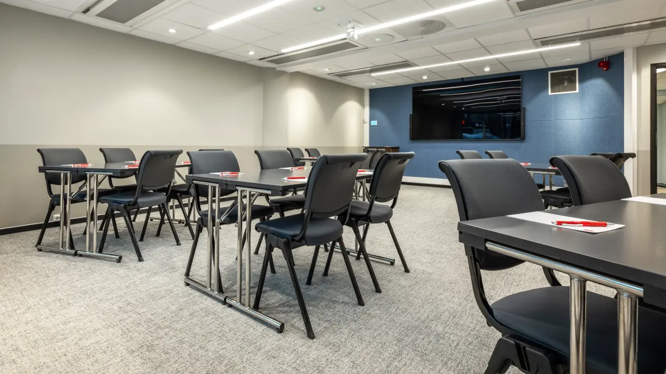 Møterom med flere gruppebord, en blå vegg i enden med TV skjerm, penn og papir lagt ut på bordene.