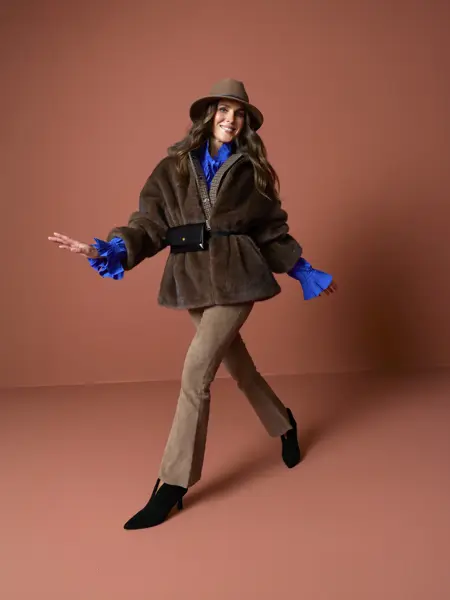 Nina i hatt, brun fuskepelsjakke, brune bukser, knallblå skjorte og støveltter.