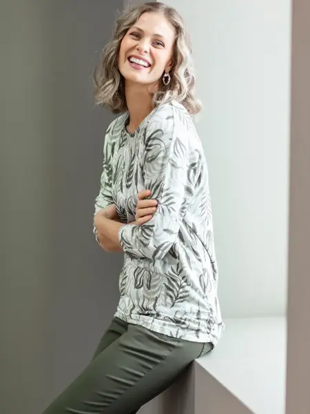 Smilende dame sitter halvveis i en vinduskarm, ser på kameraet. Har på seg grønne bukser, og lys topp med grønne blad på. 