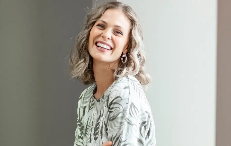 Smilende dame sitter halvveis i en vinduskarm, ser på kameraet. Har på seg grønne bukser, og lys topp med grønne blad på. 