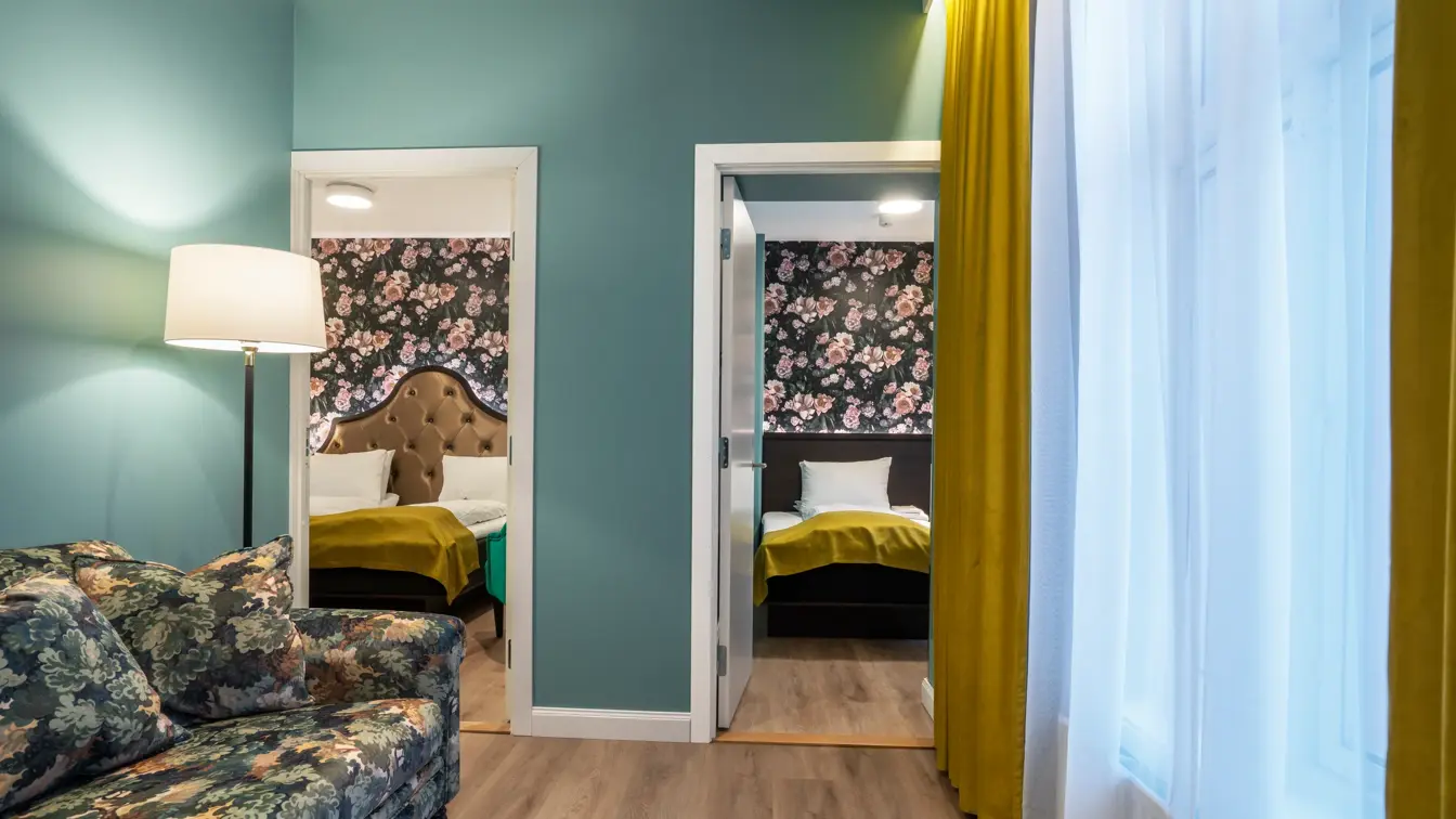 Stue med stort vindu med gule gardiner fra tak til gulv. Turkise vegger, mønstrete sofa og dør inn til soverom. I business rom på Thon Hotel Høyers i Skien.