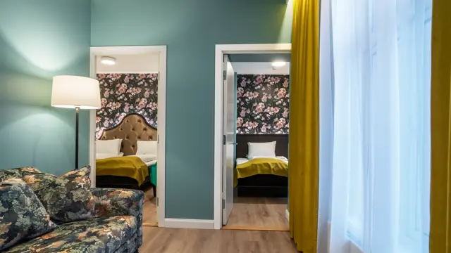 Stue med stort vindu med gule gardiner fra tak til gulv. Turkise vegger, mønstrete sofa og dør inn til soverom. I business rom på Thon Hotel Høyers i Skien.