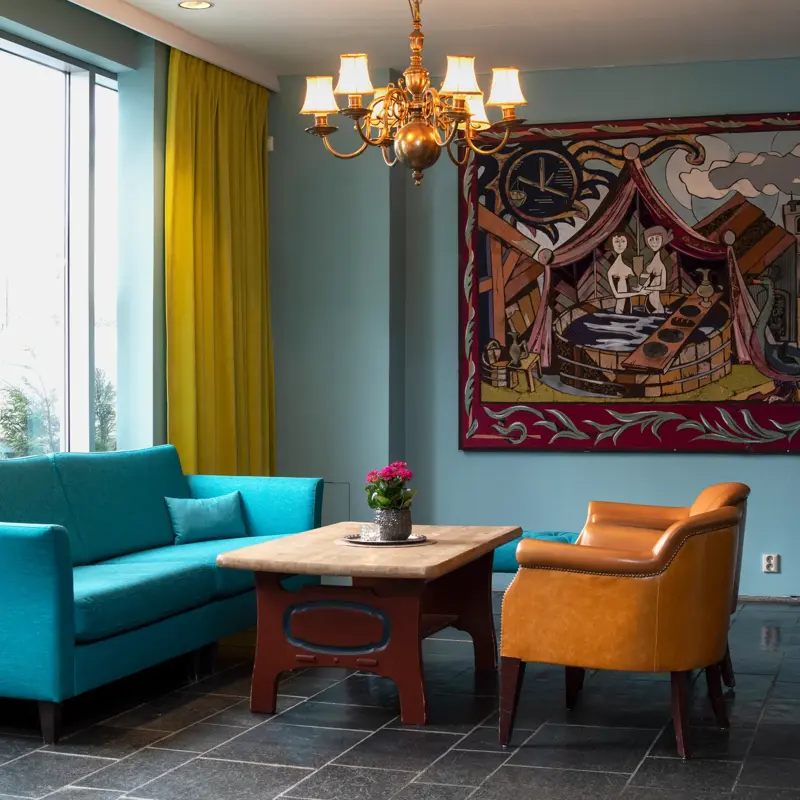 Blå sofa, to oransje stoler, flisgulver, salongbord, vindu fra gulv til tak, gule gardiner, maleri. 