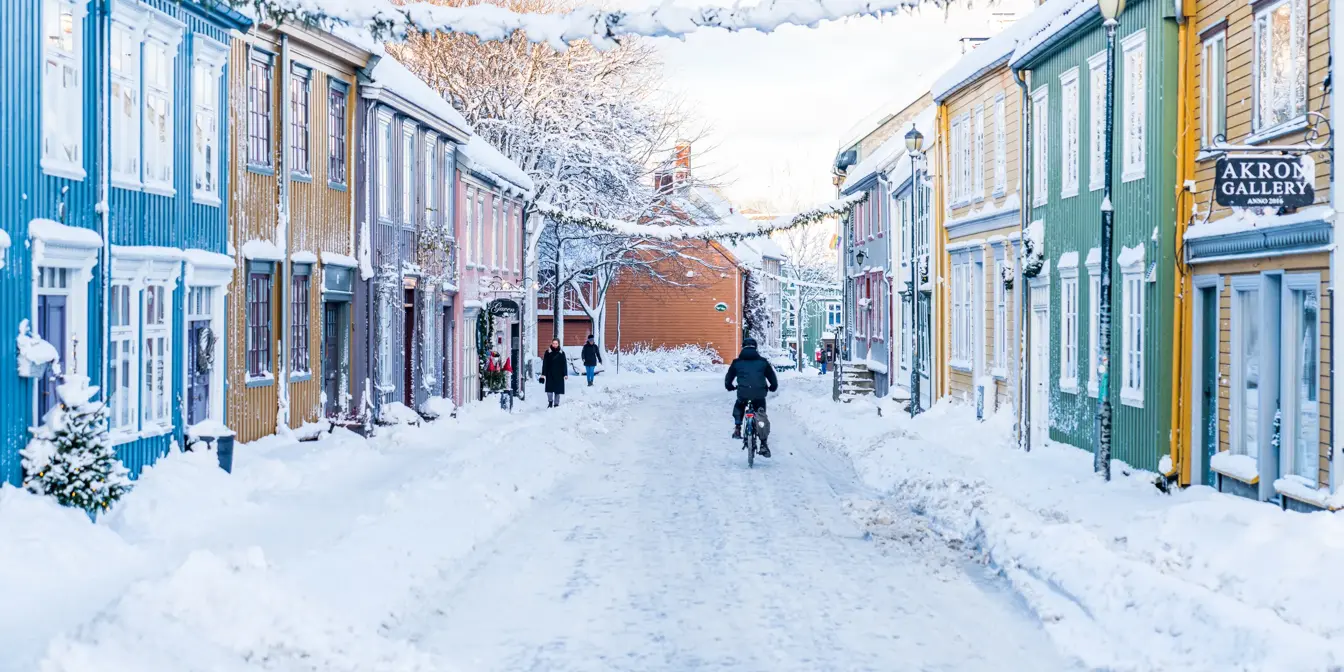 Et snødekket Bakklandet i Trondheim. Med fargerike bygg på begge sider. En syklist sykler i veien, under julepynt som heger fra den ene til den andre siden av gaten. På fortauet går to personer.