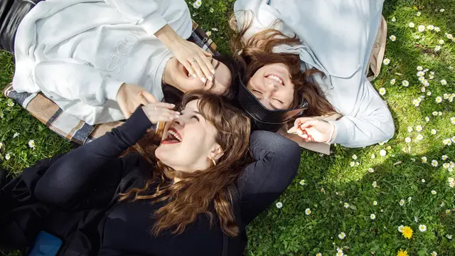 Tre damer i collegegensere ligger på blomstereng og ler 