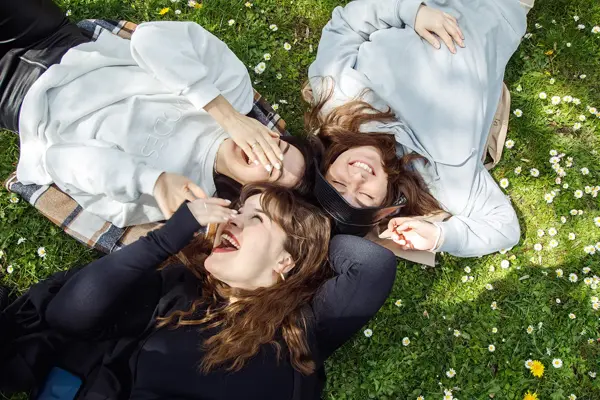 Tre damer i collegegensere ligger på blomstereng og ler 