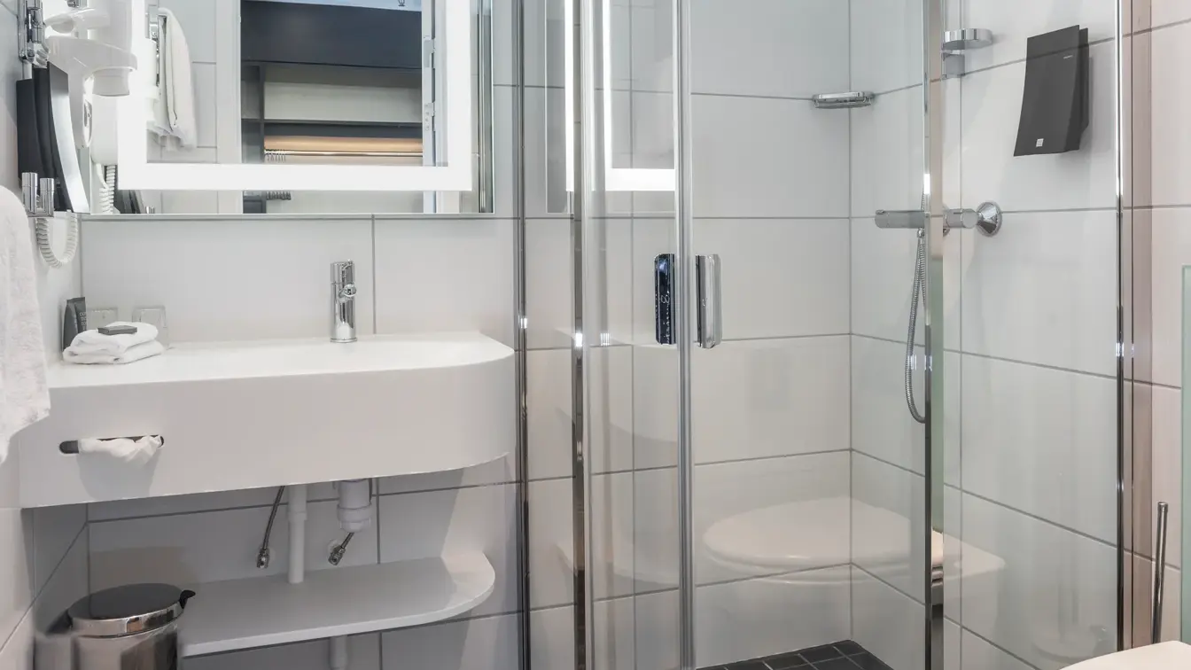 Svarte gulvfliser med hvite veggfliser, hvit porselen vask, glassdusj kabinett og et hvitt  porselens toalett.