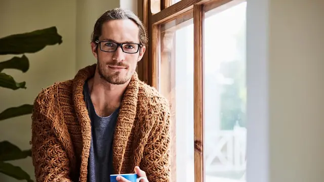 Mann med briller og brun strikkejakke som holder en kopp og sitter ved et vindu og ser i kamera