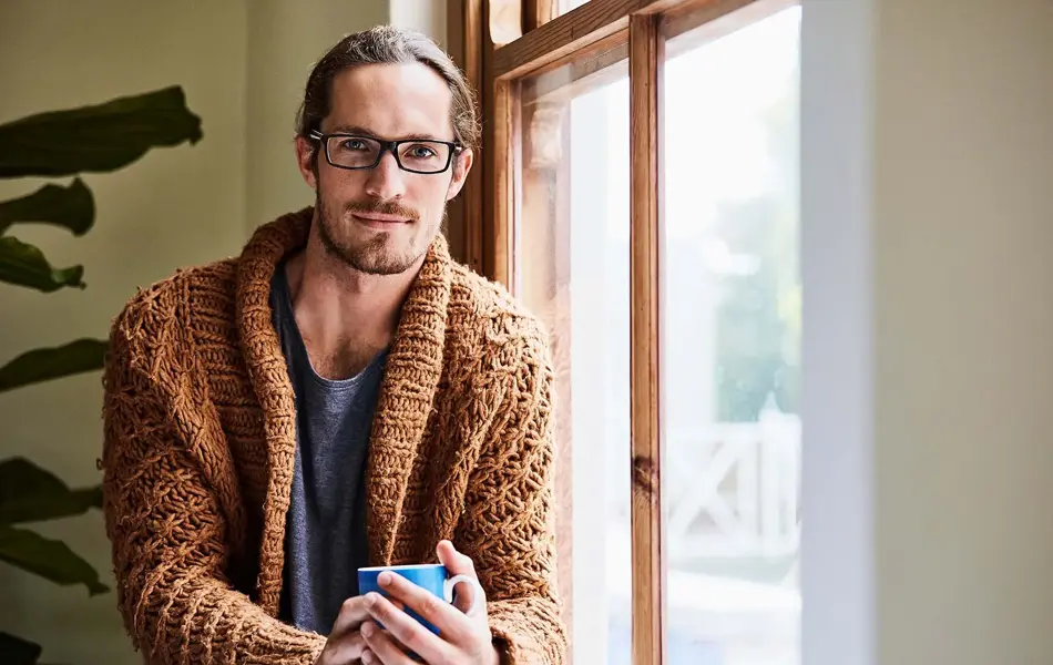 Mann med briller og brun strikkejakke som holder en kopp og sitter ved et vindu og ser i kamera
