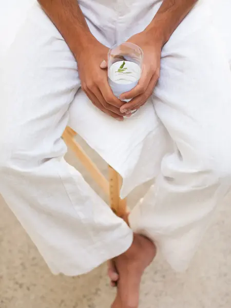 Nærbilde av manneben med hvit linbukse sittende på stol med hender som holder et vannglass 