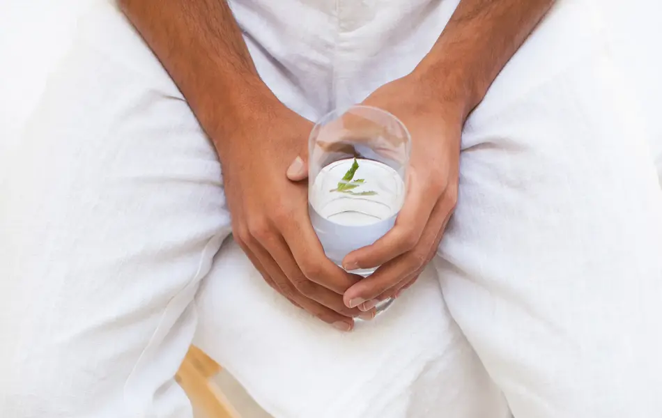 Nærbilde av manneben med hvit linbukse sittende på stol med hender som holder et vannglass 