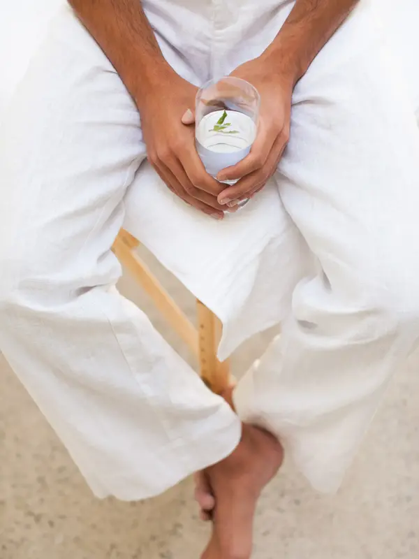 Nærbilde av manneben med hvit linbukse sittende på stol med hender som holder et vannglass 