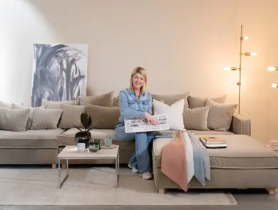 En dame som holder på med oppussing og tips om interiør trender for våren 2021. Damen har lyst halvlangt hå og har på seg en blå olaskjorte og blå olabukse. Fargene går i brunt og beige jordtoner og det er mye lilla og pastellfarger. Vårens interiør