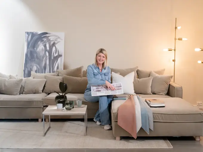 En dame som holder på med oppussing og tips om interiør trender for våren 2021. Damen har lyst halvlangt hå og har på seg en blå olaskjorte og blå olabukse. Fargene går i brunt og beige jordtoner og det er mye lilla og pastellfarger. Vårens interiør