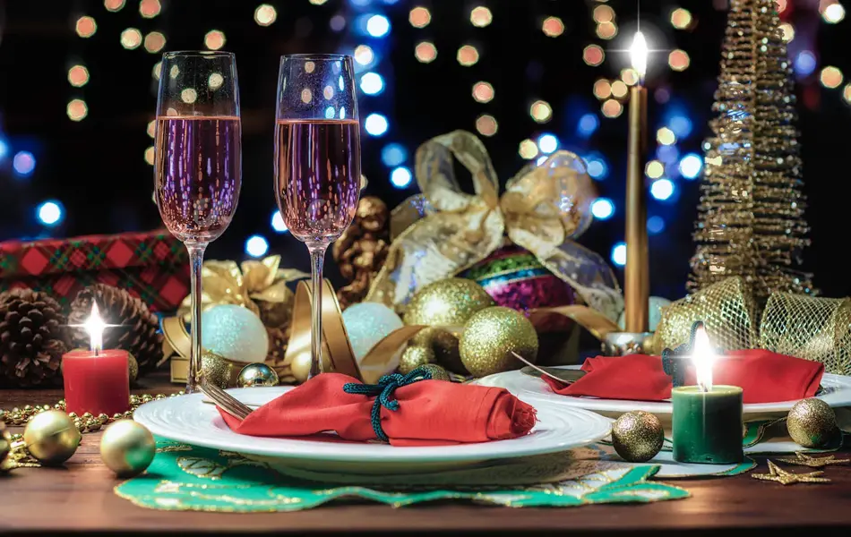 Bord som er pyntet til nyttårsfest, med røde servietter, julekuler, lys og to champagneglass. Foto til artikkel om hvordan du fikser nyttårsfest uten å slite deg helt ut.