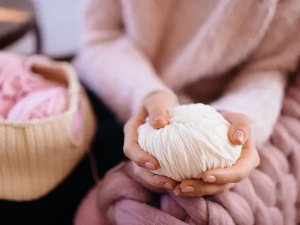 Woman in a soft knitted pink sweater sitting in chair and holding white yarn. Knitted sweater. Knitter. Heap balls of yarn on table. Fall or winter time concept. Cotton, wool, knitwear