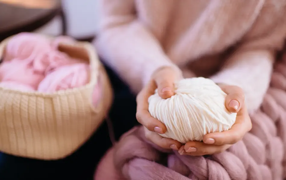 Woman in a soft knitted pink sweater sitting in chair and holding white yarn. Knitted sweater. Knitter. Heap balls of yarn on table. Fall or winter time concept. Cotton, wool, knitwear