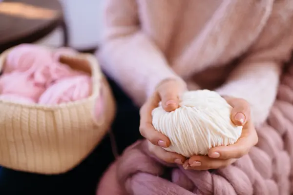 Woman in a soft knitted pink sweater sitting in chair and holding white yarn. Knitted sweater. Knitter. Heap balls of yarn on table. Fall or winter time concept. Cotton, wool, knitwear